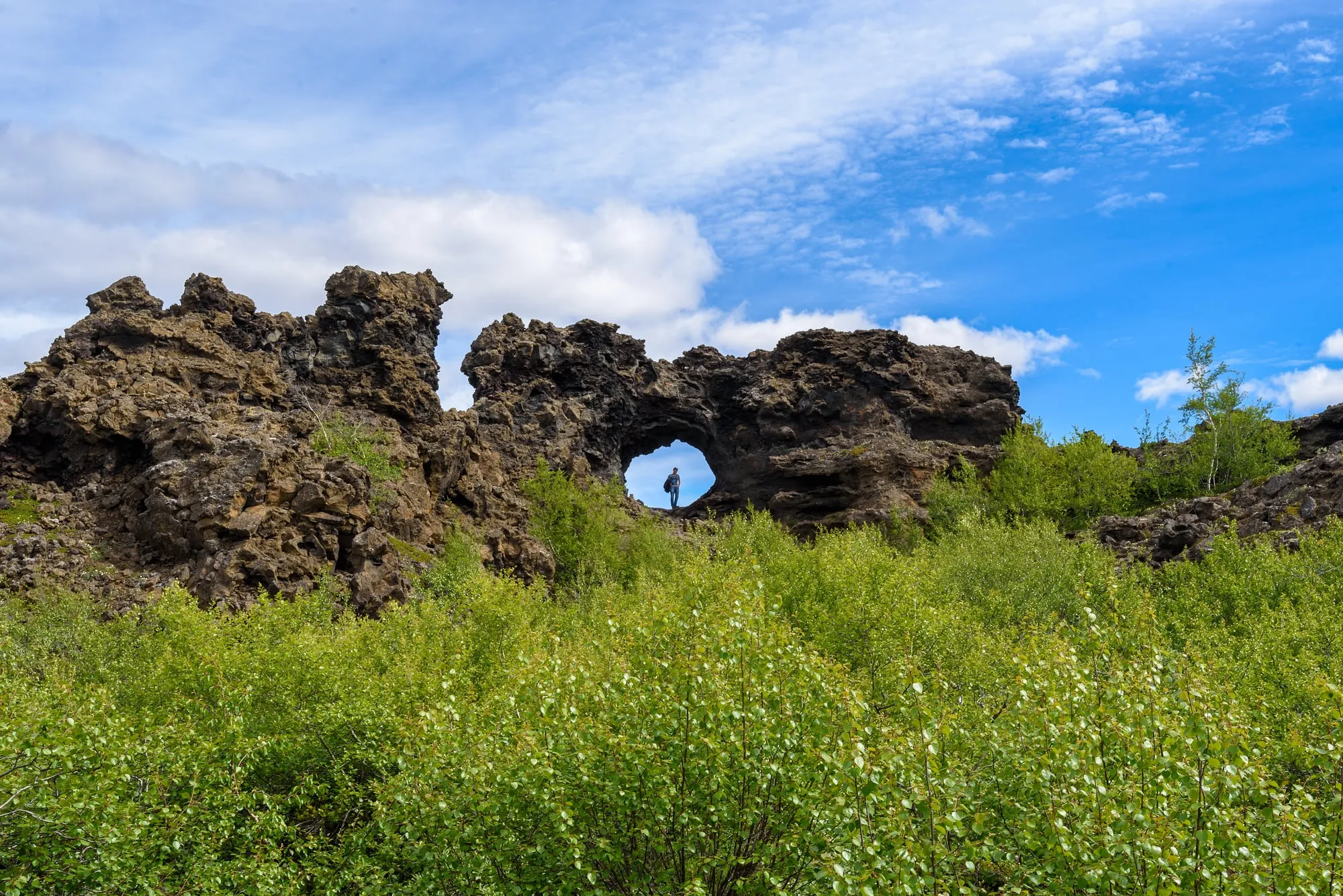 Lawowa formacja z naturalnym łukiem w Dimmuborgir, wokół zielone zarośla i błękitne niebo
