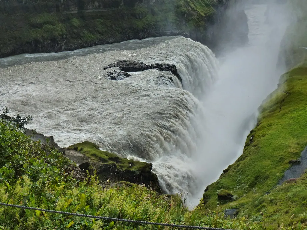 Dolna kaskada Gullfoss wpadająca do wąskiego kanionu, z boku zielone, porośnięte mchem zbocza i lina zabezpieczająca ścieżkę