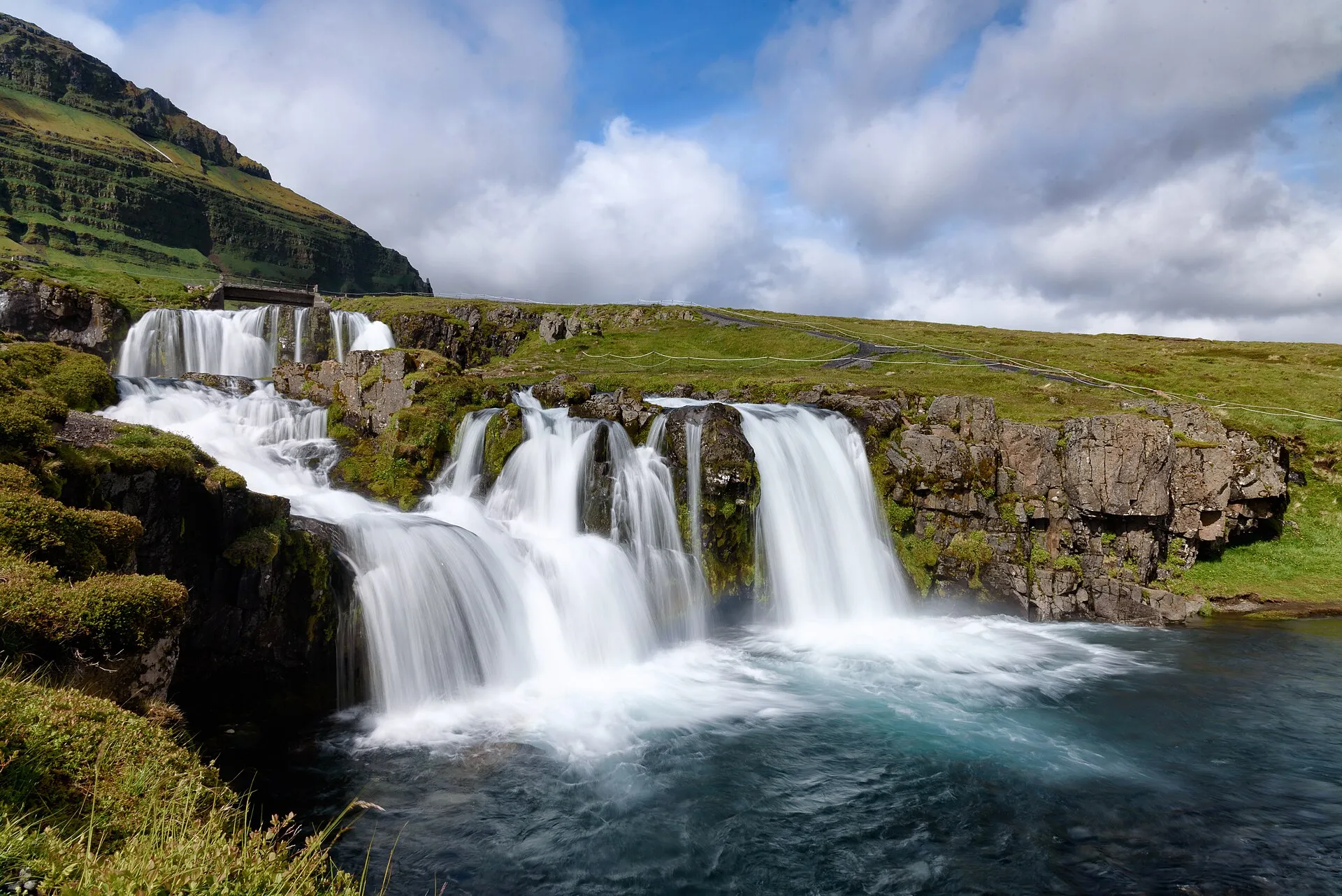 Kaskady wodospadu Kirkjufellsfoss wpadające do turkusowego oczka wody, w tle zielone zbocze Kirkjufell