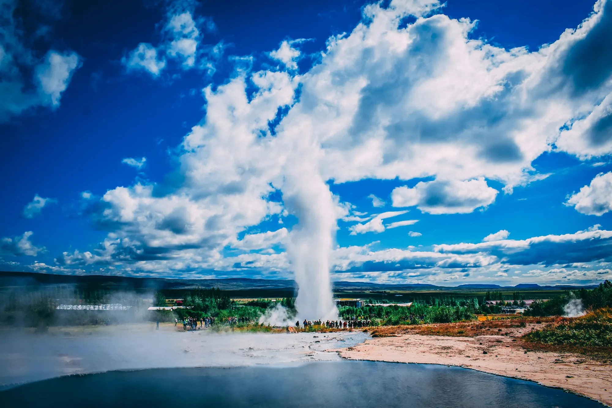 Gejzer Strokkur wyrzucający słup wody i pary, w tle turyści i zielona dolina