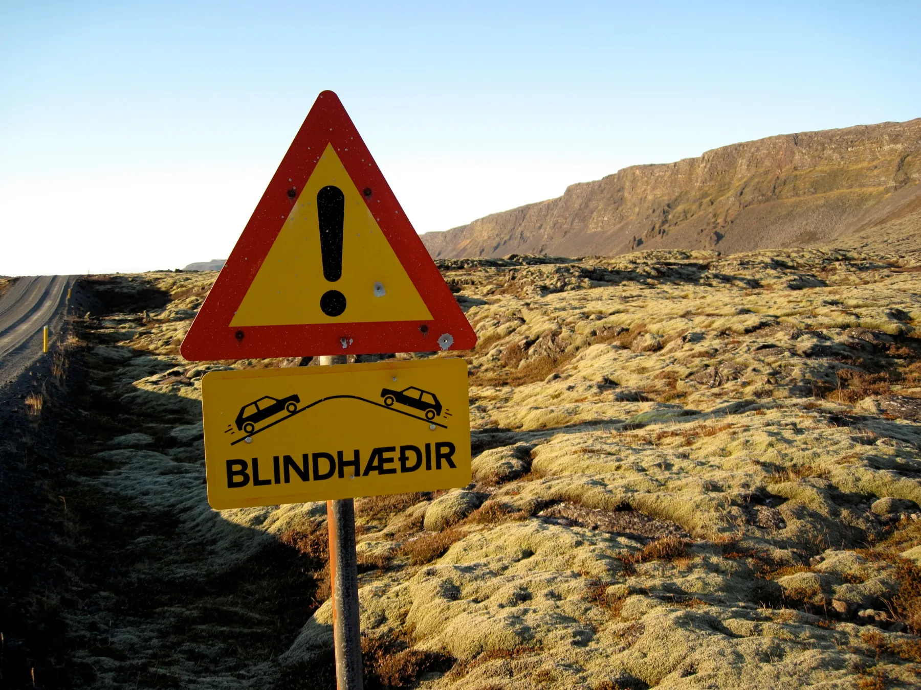 Islandzki znak ostrzegawczy Blindhæðir przy szutrowej drodze, w tle porośnięte mchem pole lawy i skalne zbocze
