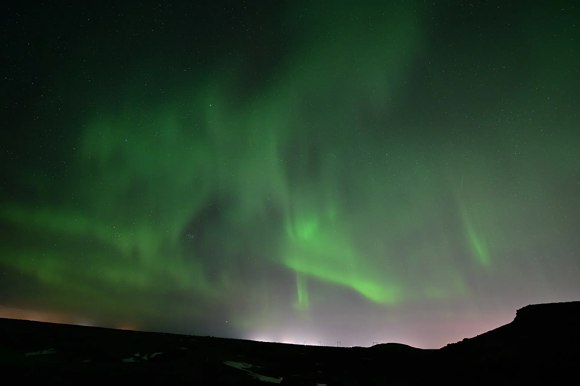 Zorza polarna nad wzgórzami, na horyzoncie poświata świateł Reykjavíku